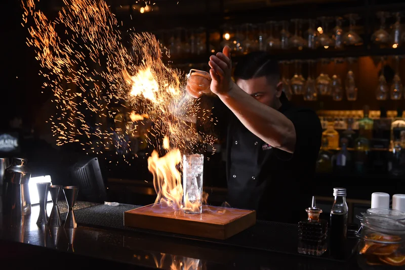Mixologist lighting a flaming cocktail serve, sparks rising over the bar